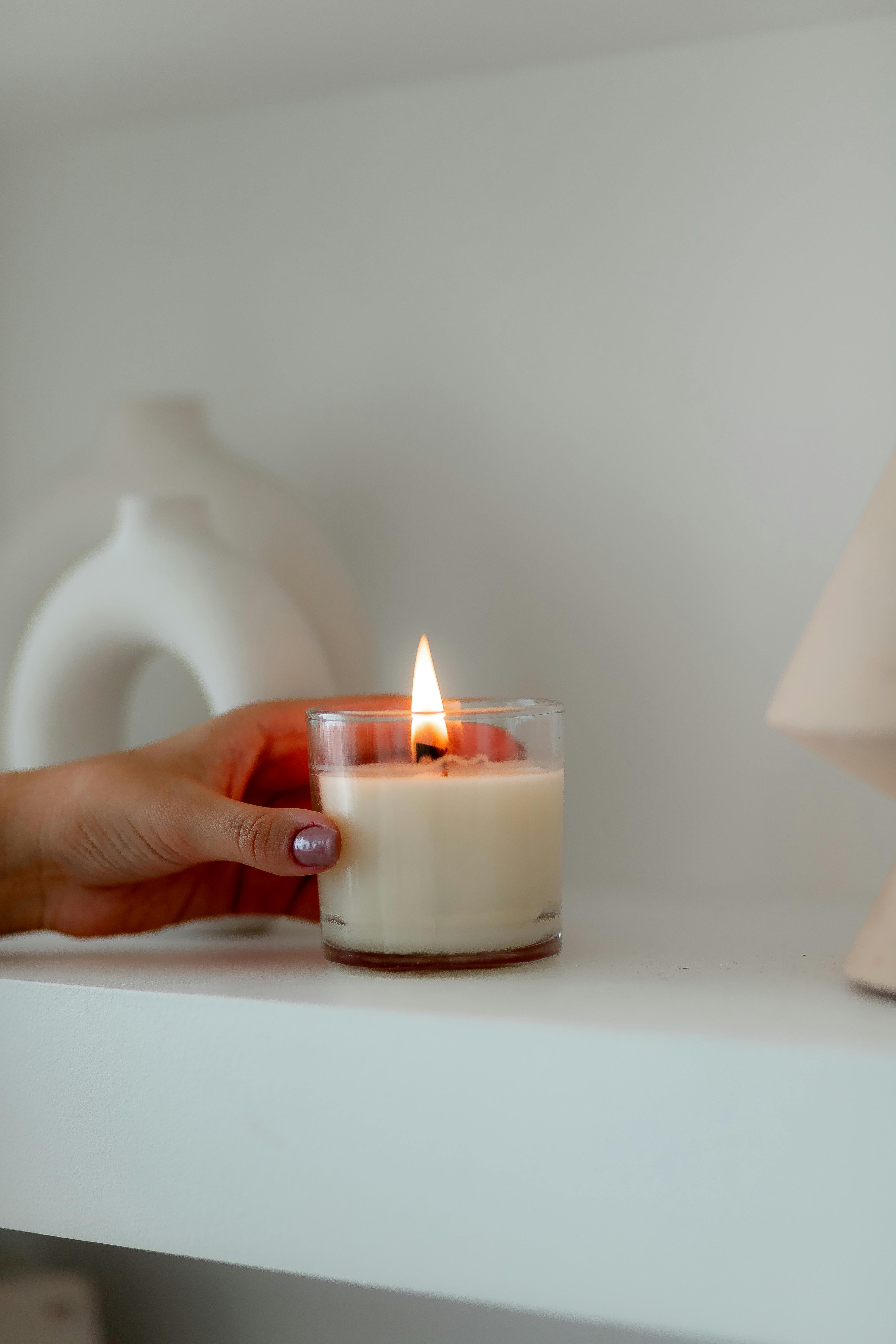 A hand with painted nails lights a white candle in a minimalist indoor setting.
