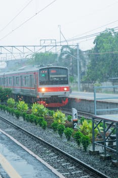 A commuter train pulling into a station during a rainy day with lush greenery nearby.