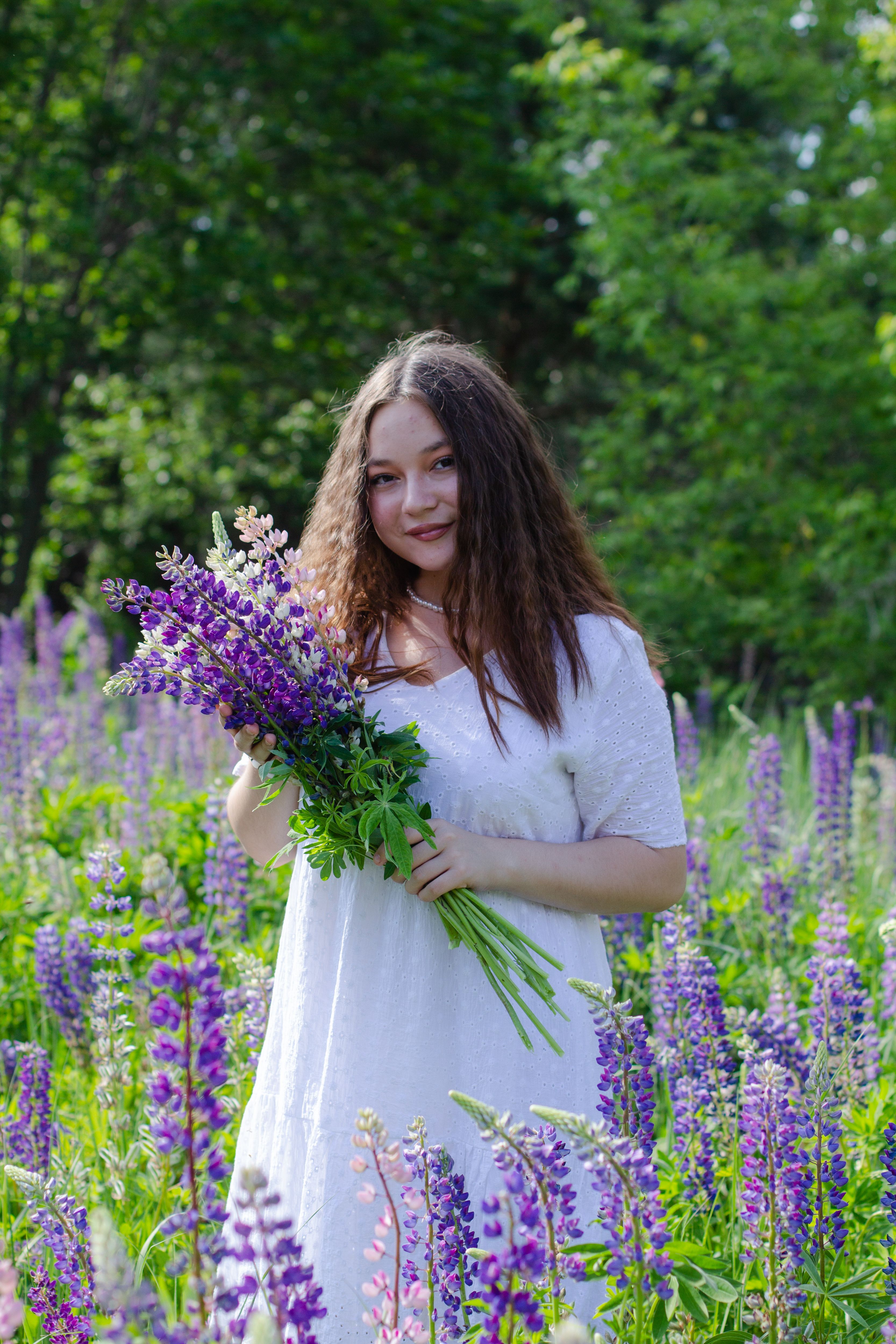 A young woman in a white dress stands in a vibrant meadow holding a bouquet of lupine flowers.