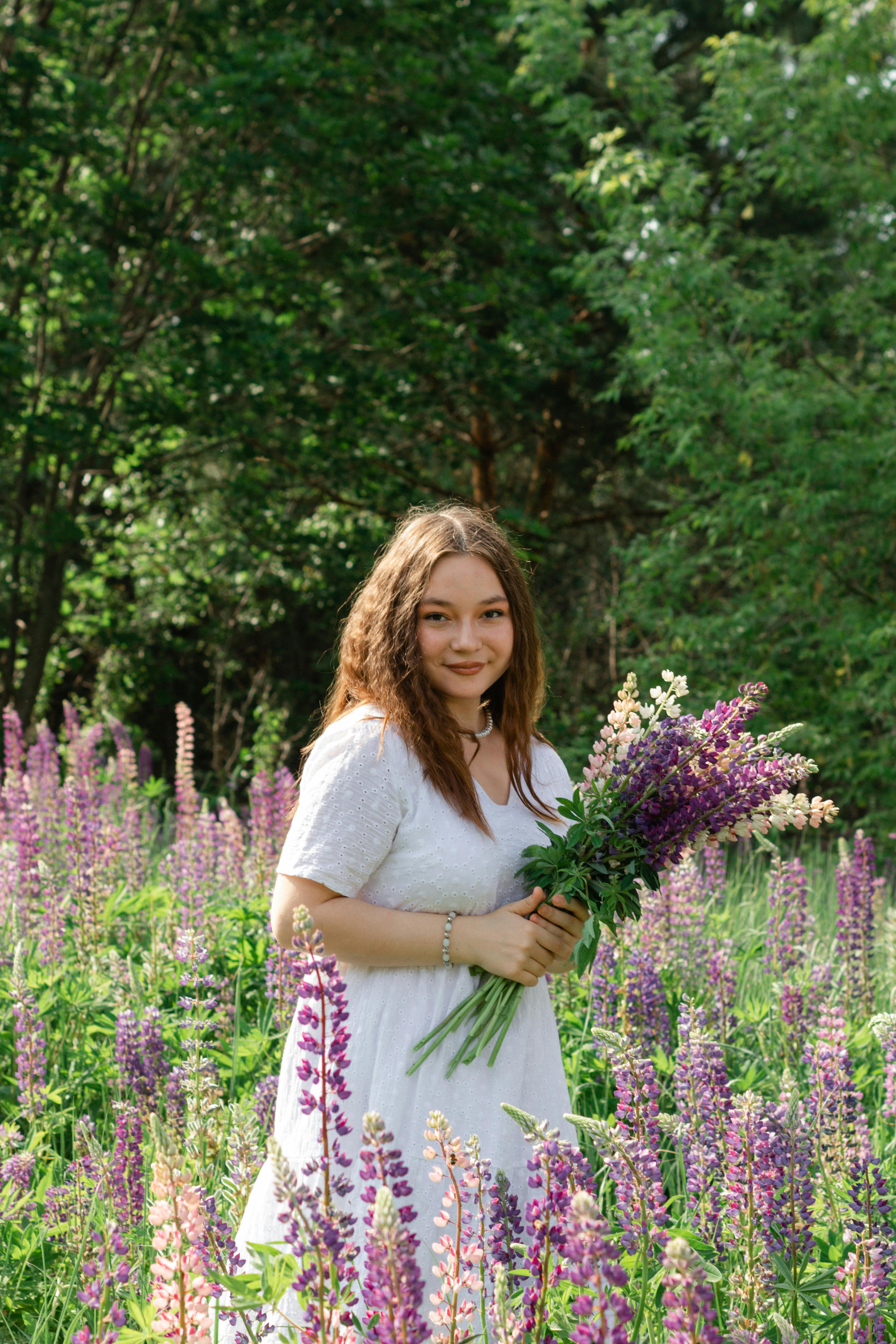 A woman in a white dress stands in a field of blooming lupines, holding a bouquet, surrounded by vibrant greenery.