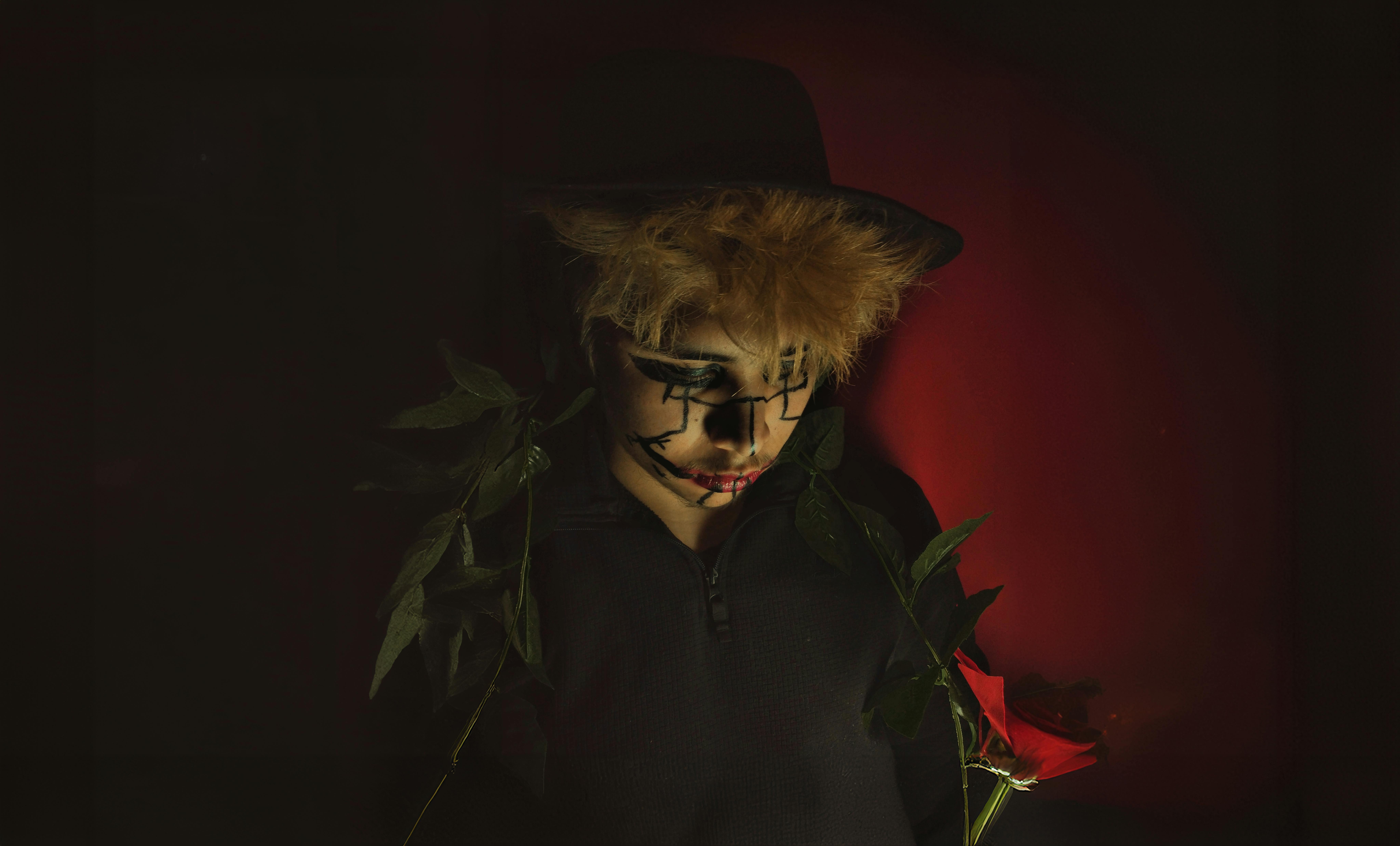 Ücretsiz Loş ışıkta, korkuluktan esinlenerek boyanmış yüze sahip, kırmızı bir gül tutan bir kişi. Stok Fotoğraflar