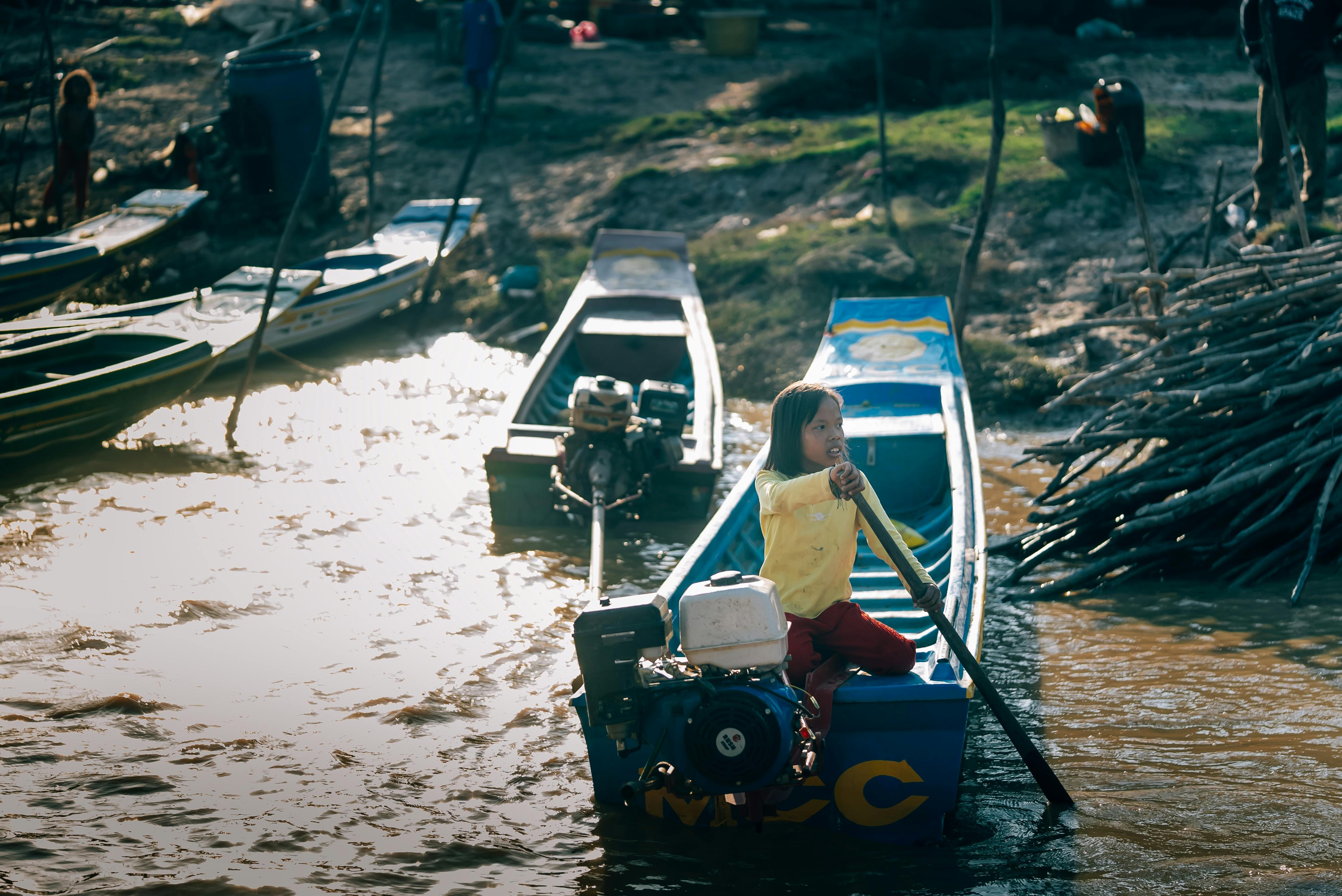무료 한 어린 소녀가 햇살이 비치는 강에서 작은 모터보트를 저으며 자연에 둘러싸여 있다. 스톡 사진