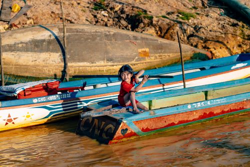 Free A young child joyfully paddles a colorful wooden boat in a serene river setting. Stock Photo