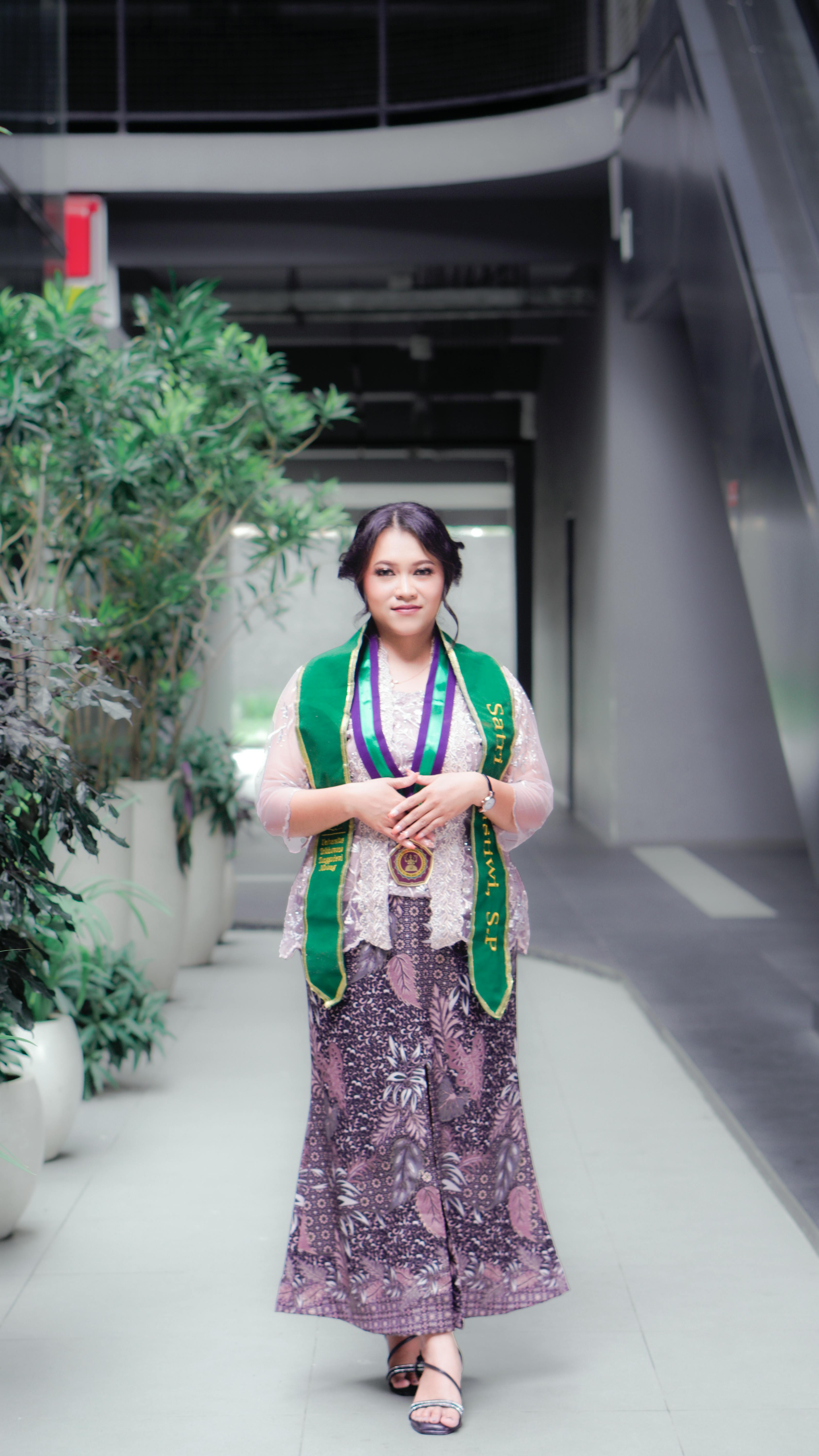 A proud graduate wearing a traditional dress and green sash in a contemporary hallway.