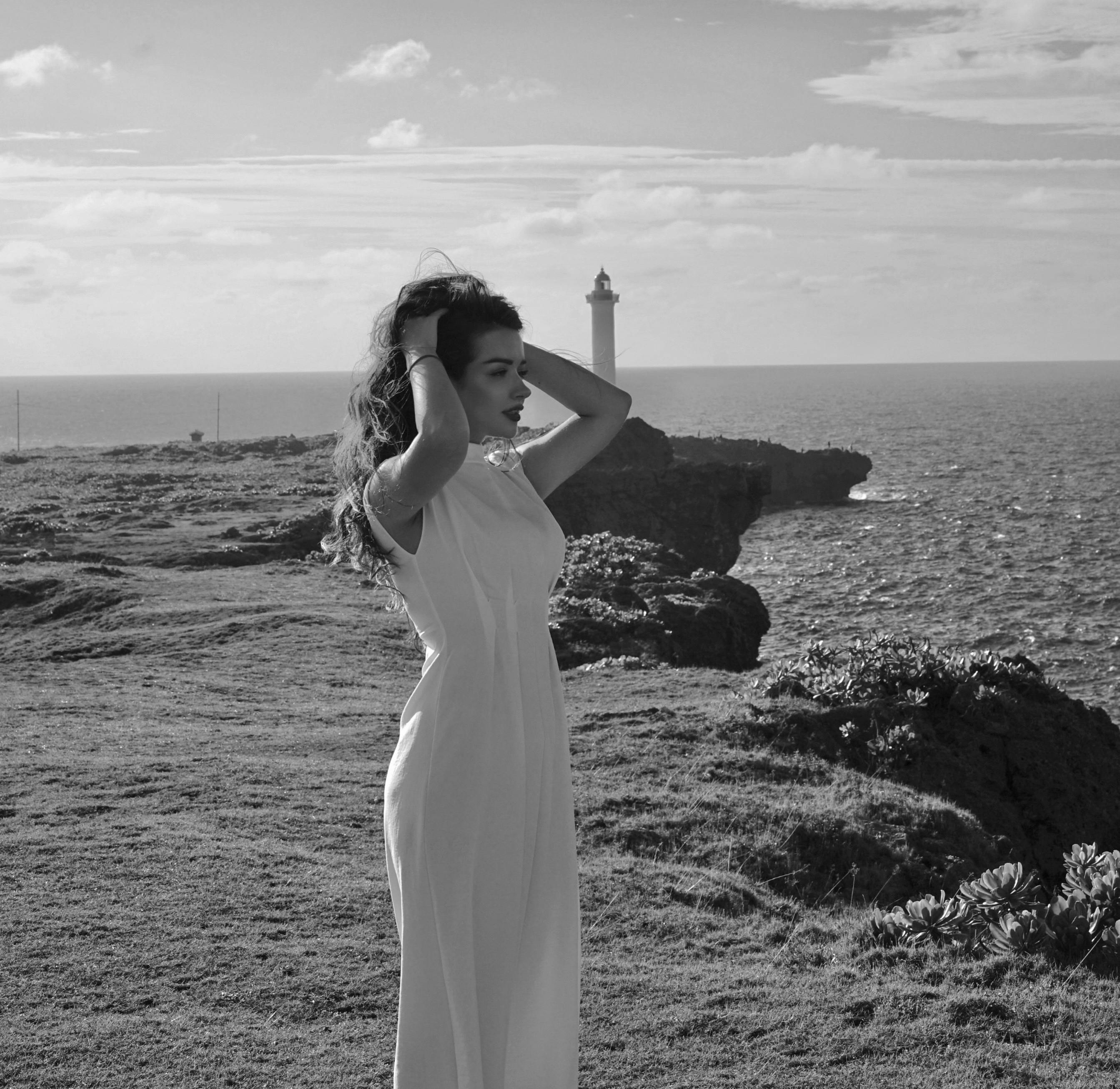 Elegant woman in a white dress standing near a cliff with a lighthouse in the background.