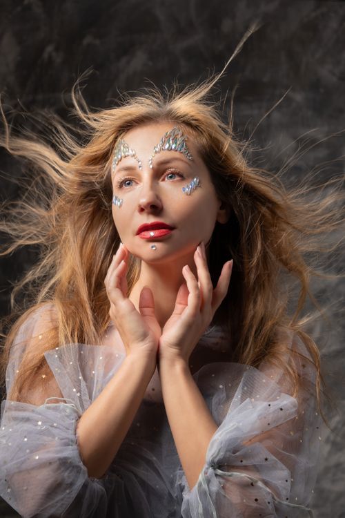Free A striking portrait of a woman with glittering face art and flowing hair in a dreamy setting. Stock Photo