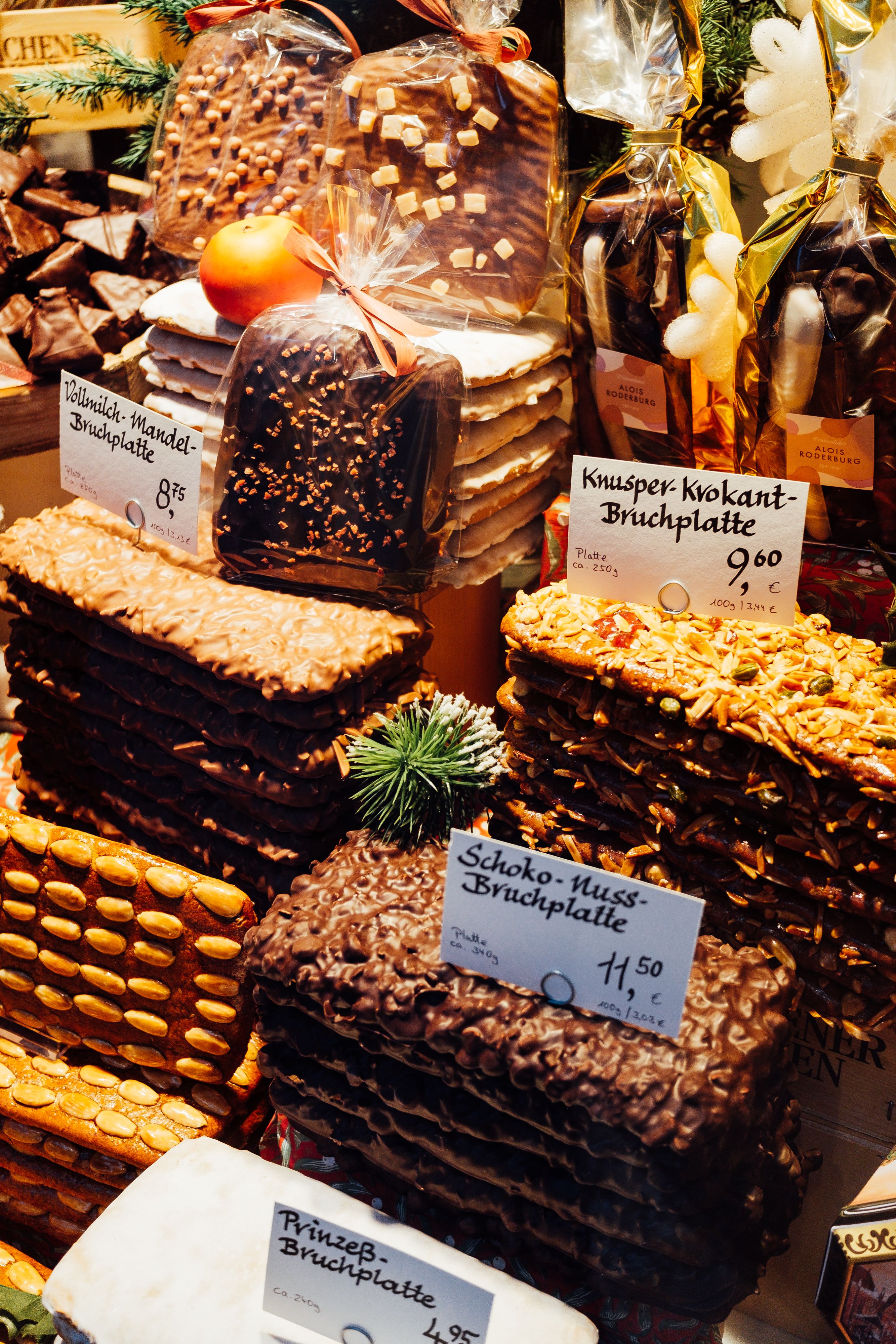 Gestapelte Lebkuchen-Bruchplatten mit Mandeln und Schokolade an einem Marktstand, davor handgeschriebene Preisschilder