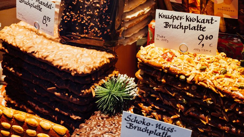 Gestapelte Lebkuchen-Bruchplatten mit Mandeln und Schokolade an einem Marktstand, davor handgeschriebene Preisschilder