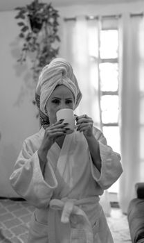 Woman in bathrobe sipping coffee, relaxing at home with natural light.