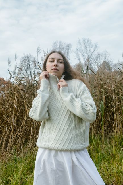Woman in a white cable knit sweater in a natural outdoor setting, exuding warm, cozy vibes.