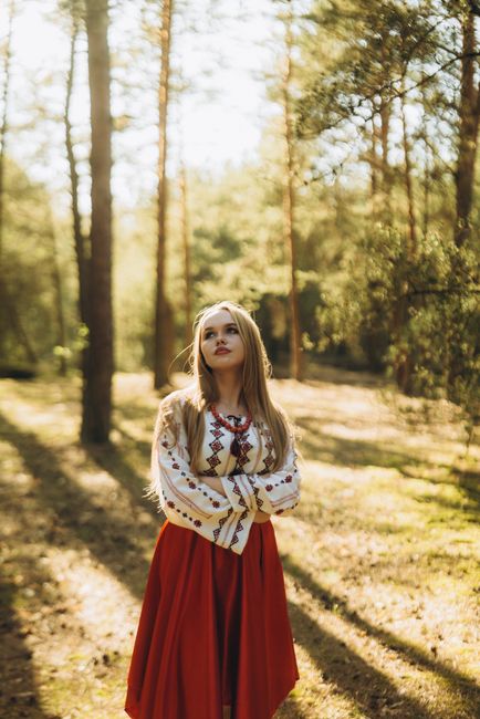 Woman in embroidered dress stands in sunlit forest, cultural attire highlights traditional elegance.