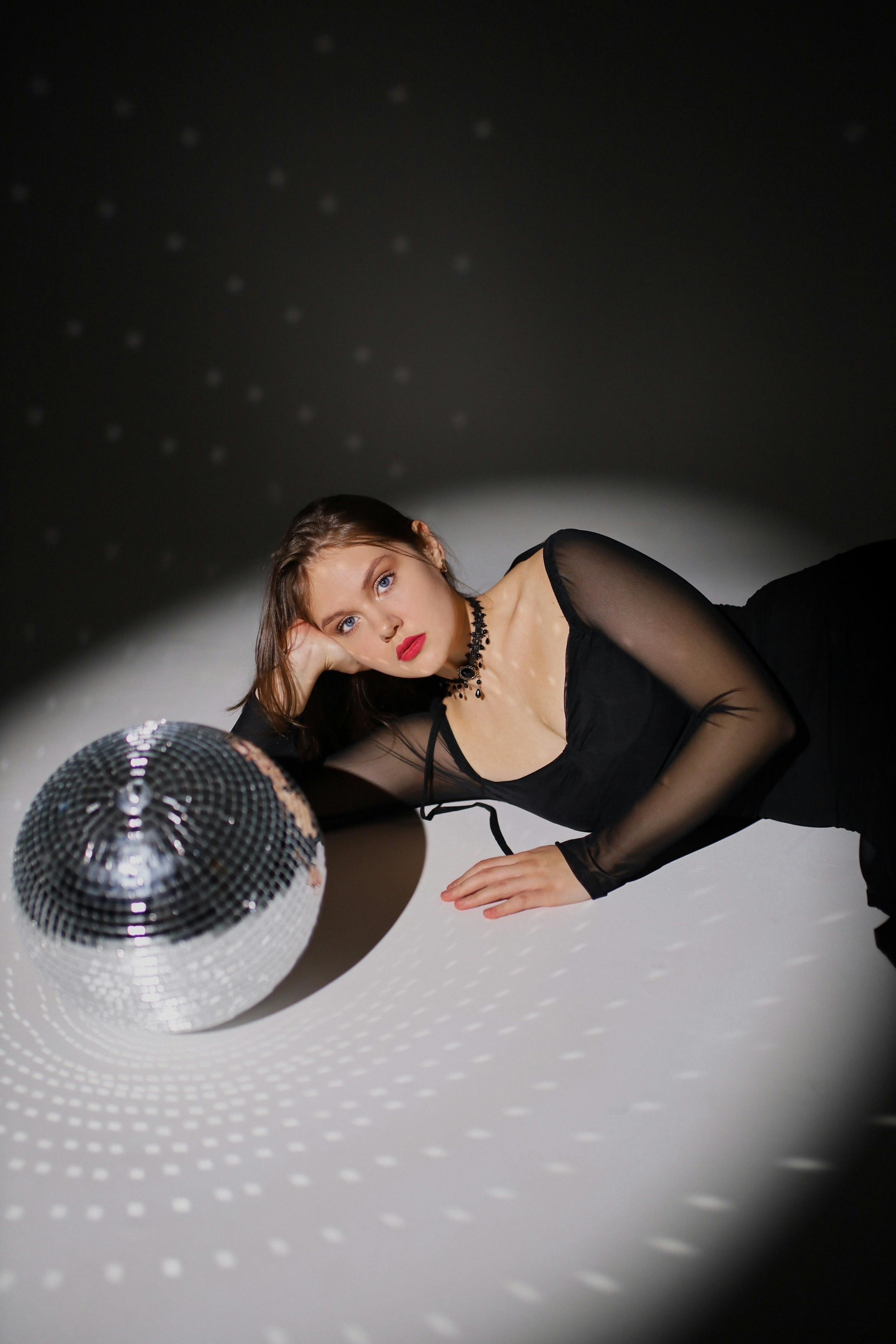 Elegant woman in black dress beside disco ball, under a spotlight.