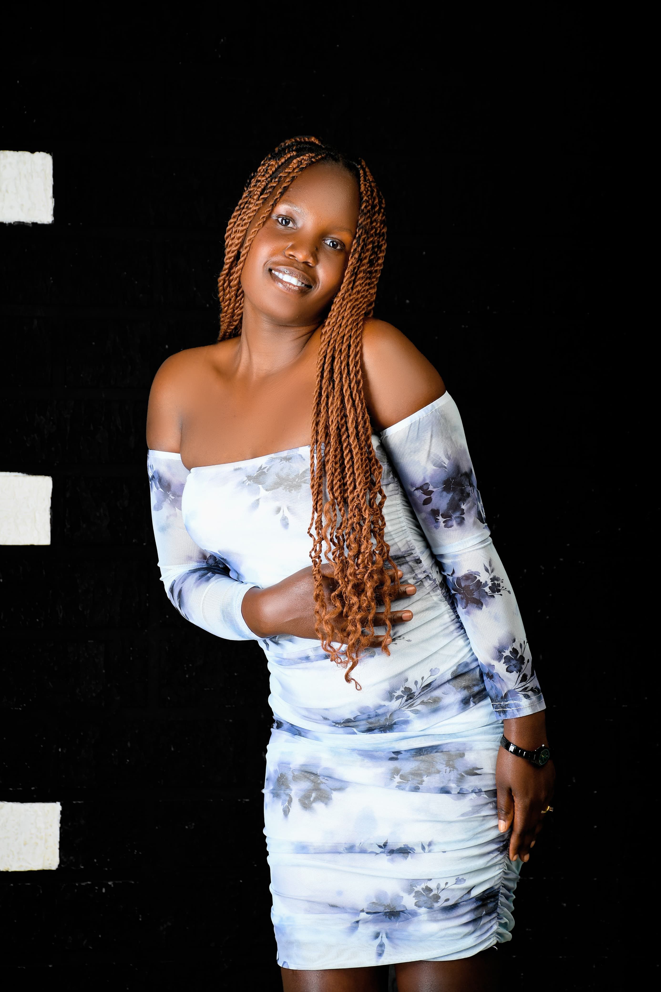 Charming woman with long braids posing in a stylish floral dress against a black backdrop.