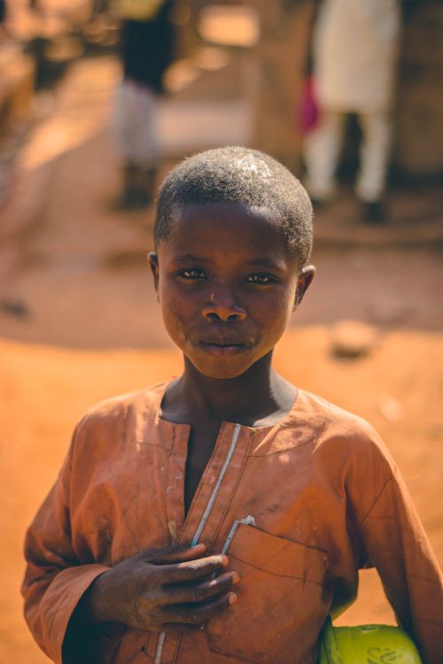 Free A young boy stands in a sunlit village, looking directly at the camera. Stock Photo