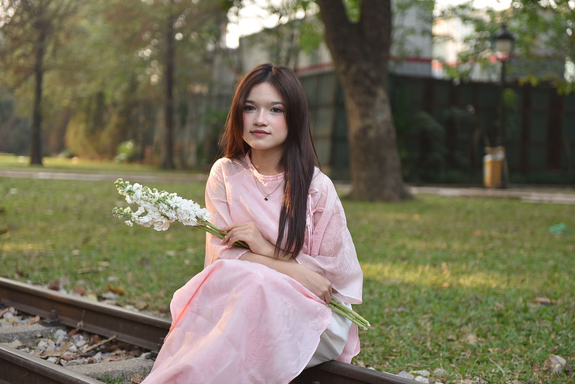 A young woman sits on railway tracks holding white flowers, outdoors.