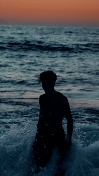 Silhouetted figure standing in ocean waves during a colorful Moroccan beach sunset.