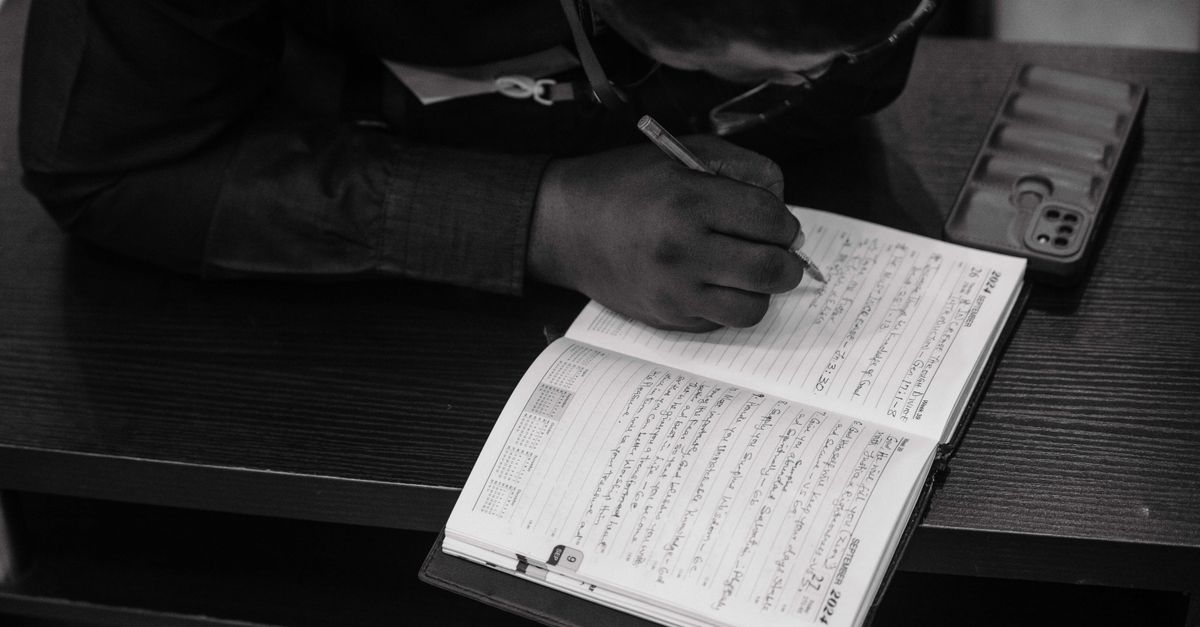 person writing in notebook with Bible open on lap during a church service