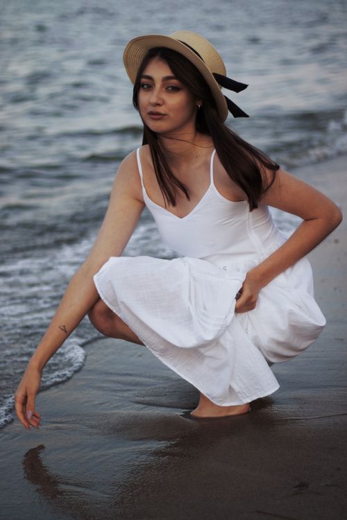 Free Graceful woman in a white dress and hat crouching by the sea shore. Stock Photo