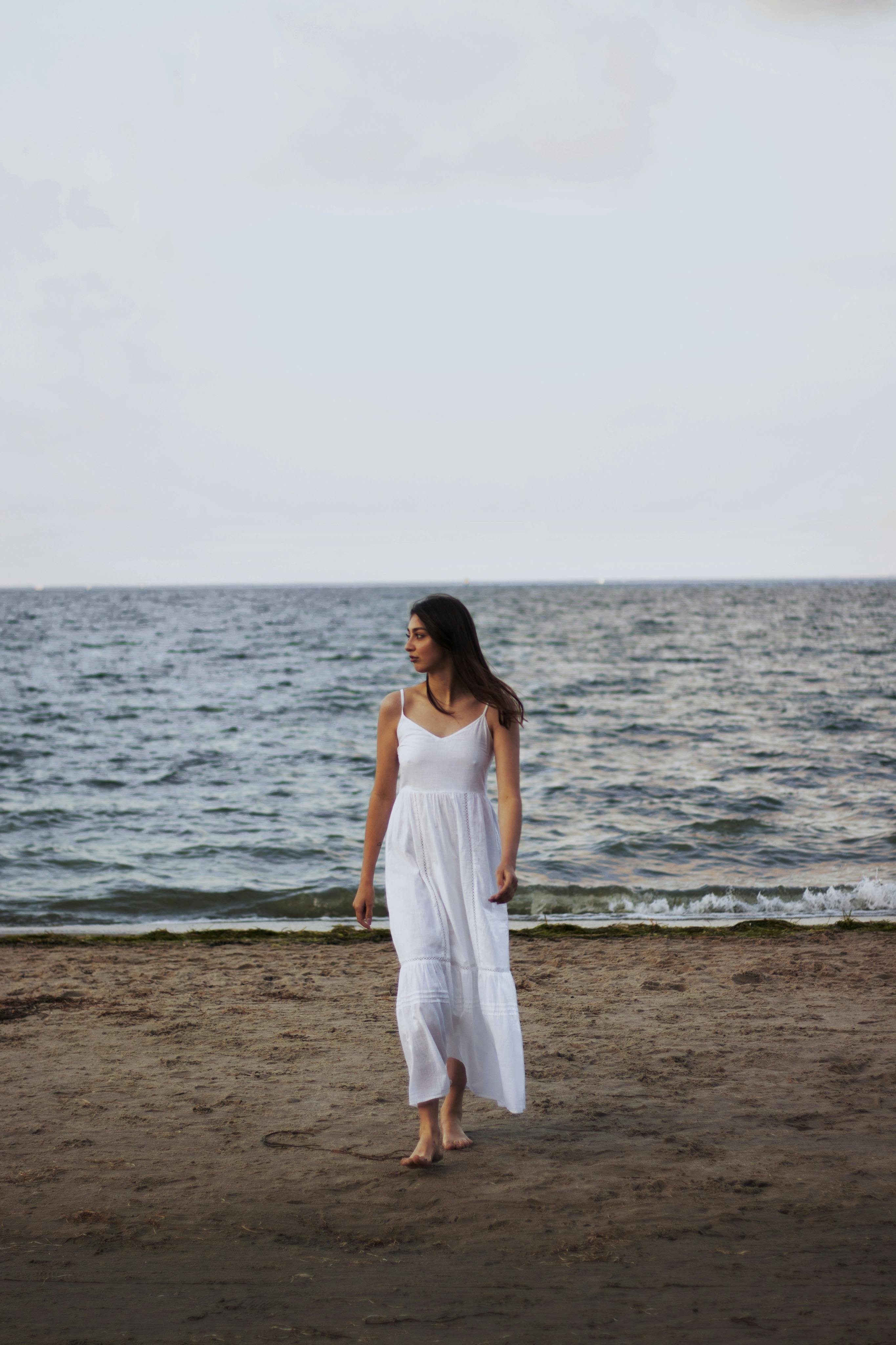 Woman in white dress walking barefoot along a peaceful beach during sunset.