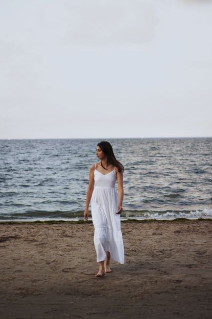 Woman in white dress walking barefoot along a peaceful beach during sunset.