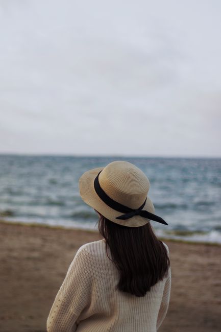 A woman in a straw hat stands at the beach, looking out at the calm ocean horizon.