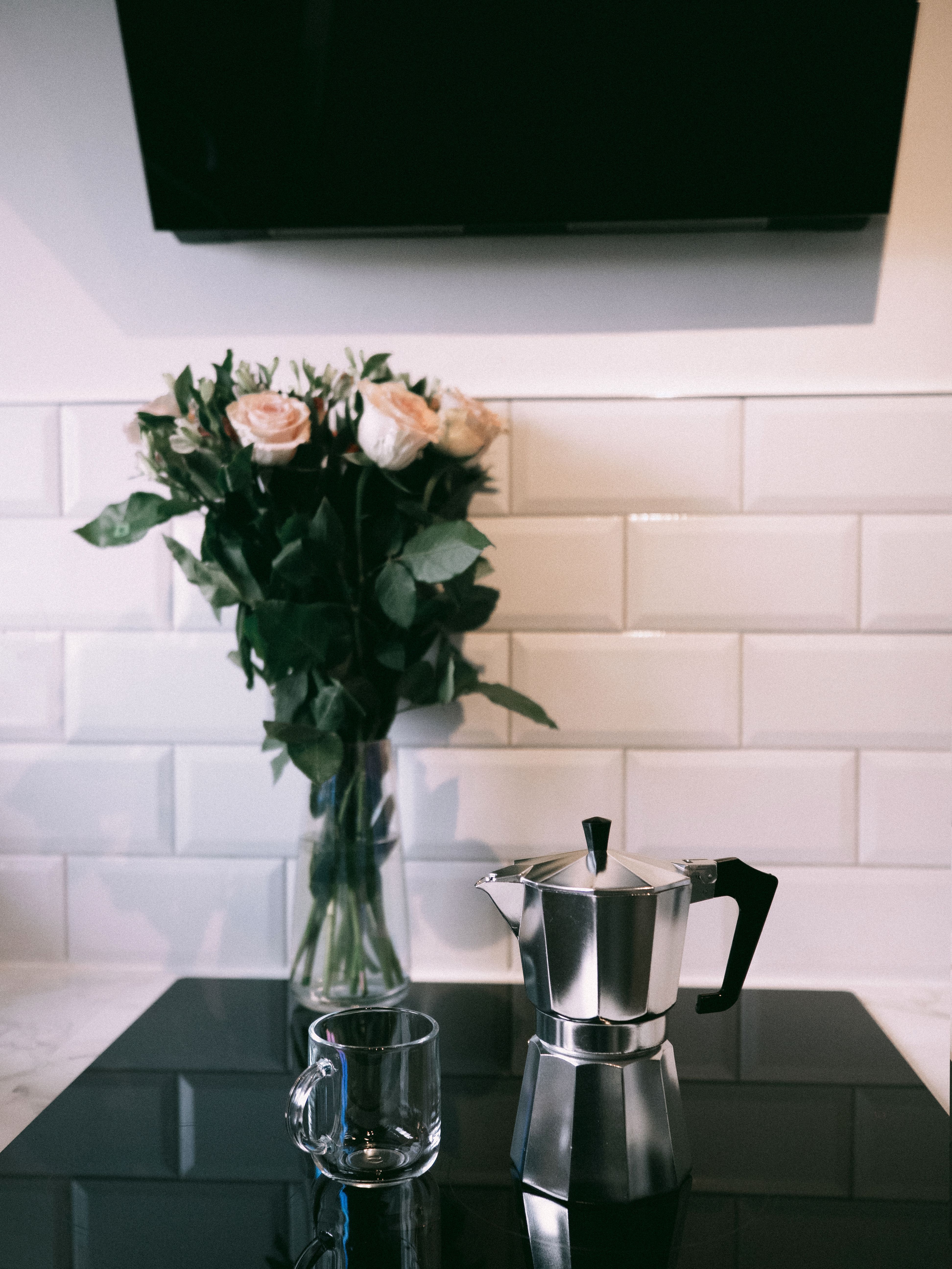 A stylish kitchen scene with a moka pot, glass mug, and vase of roses on a reflective surface.