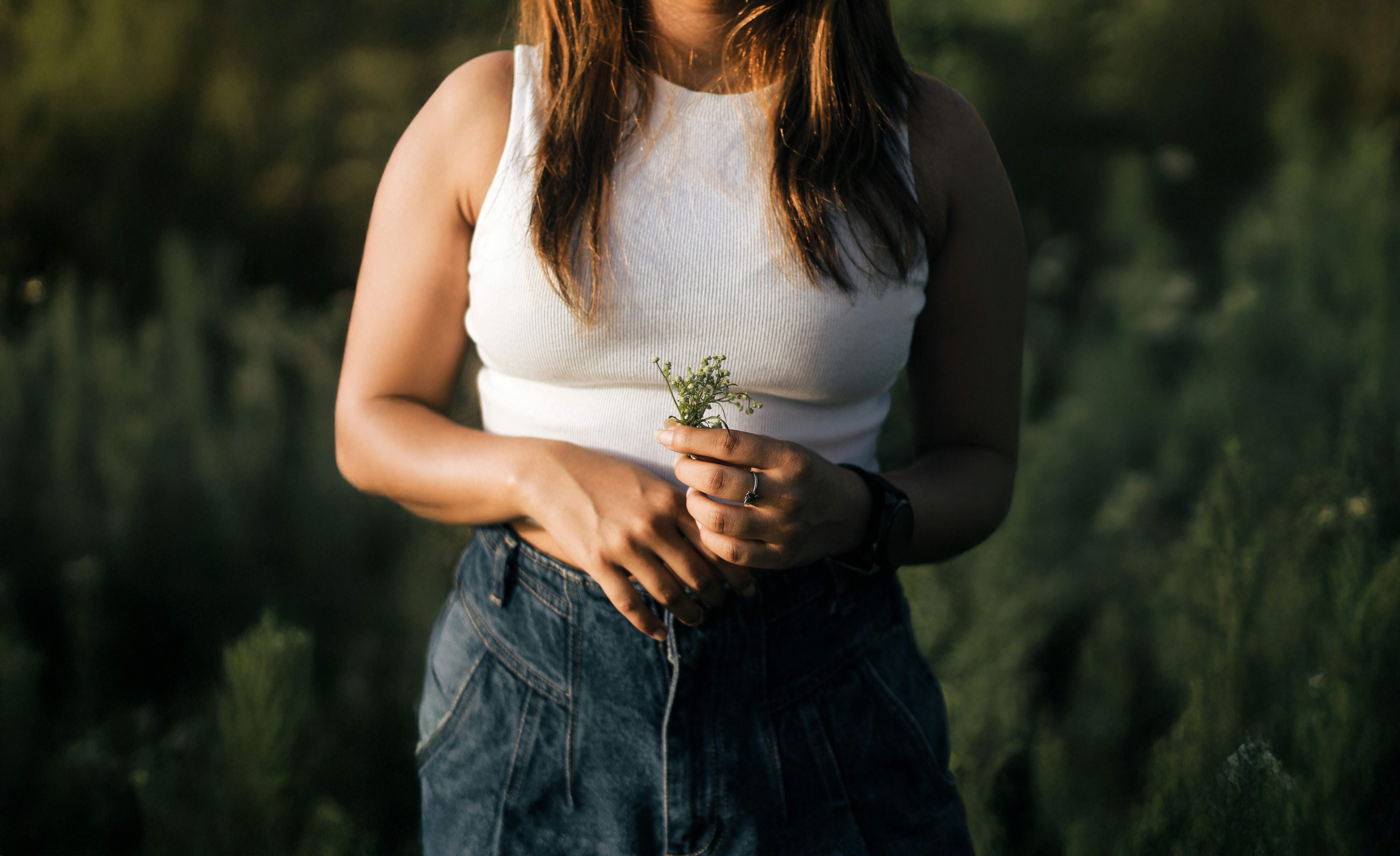 A woman in a white tank top and jeans holds a small wildflower in a sunny field.