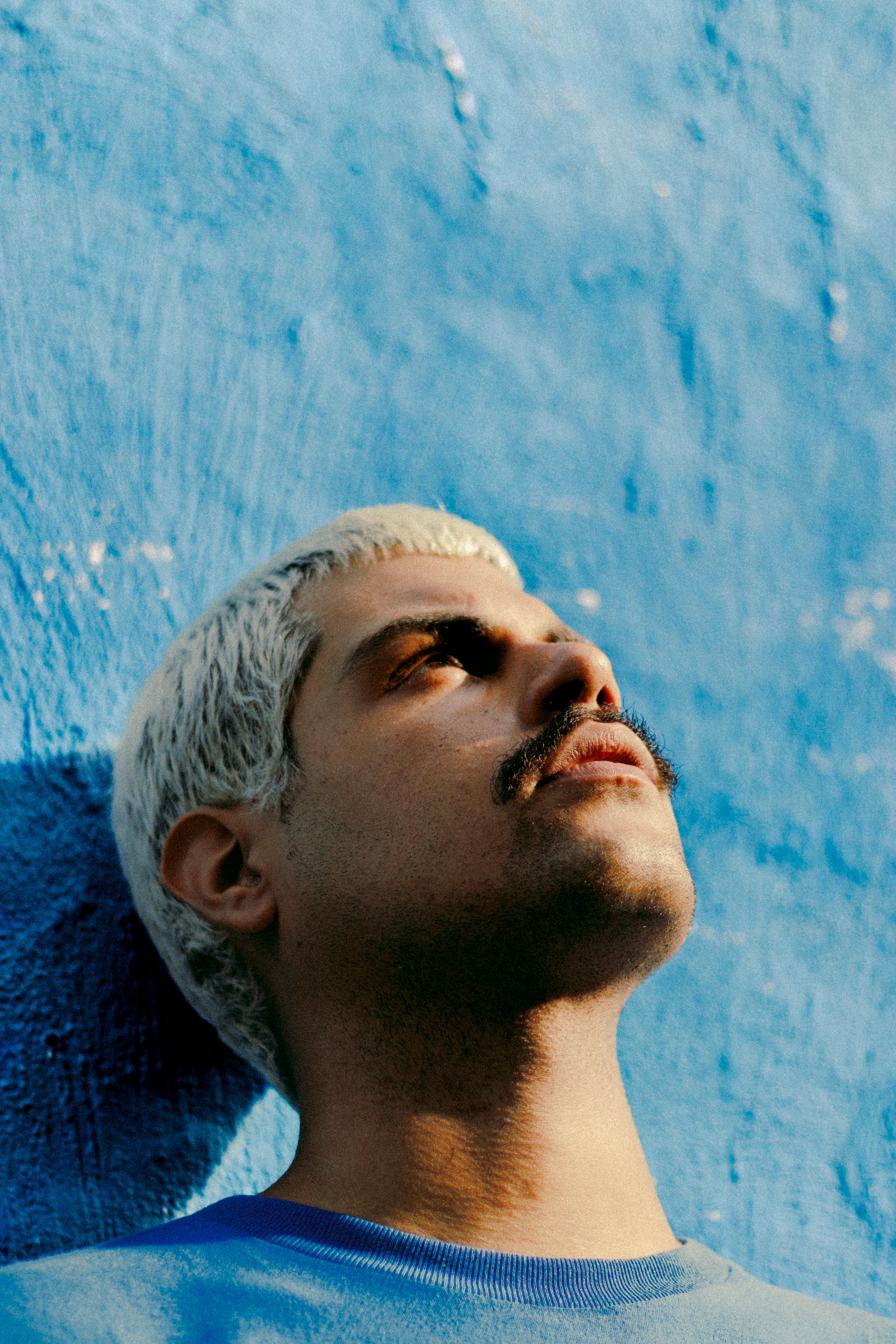 A man with platinum hair gazes up, leaning against a bright blue wall.