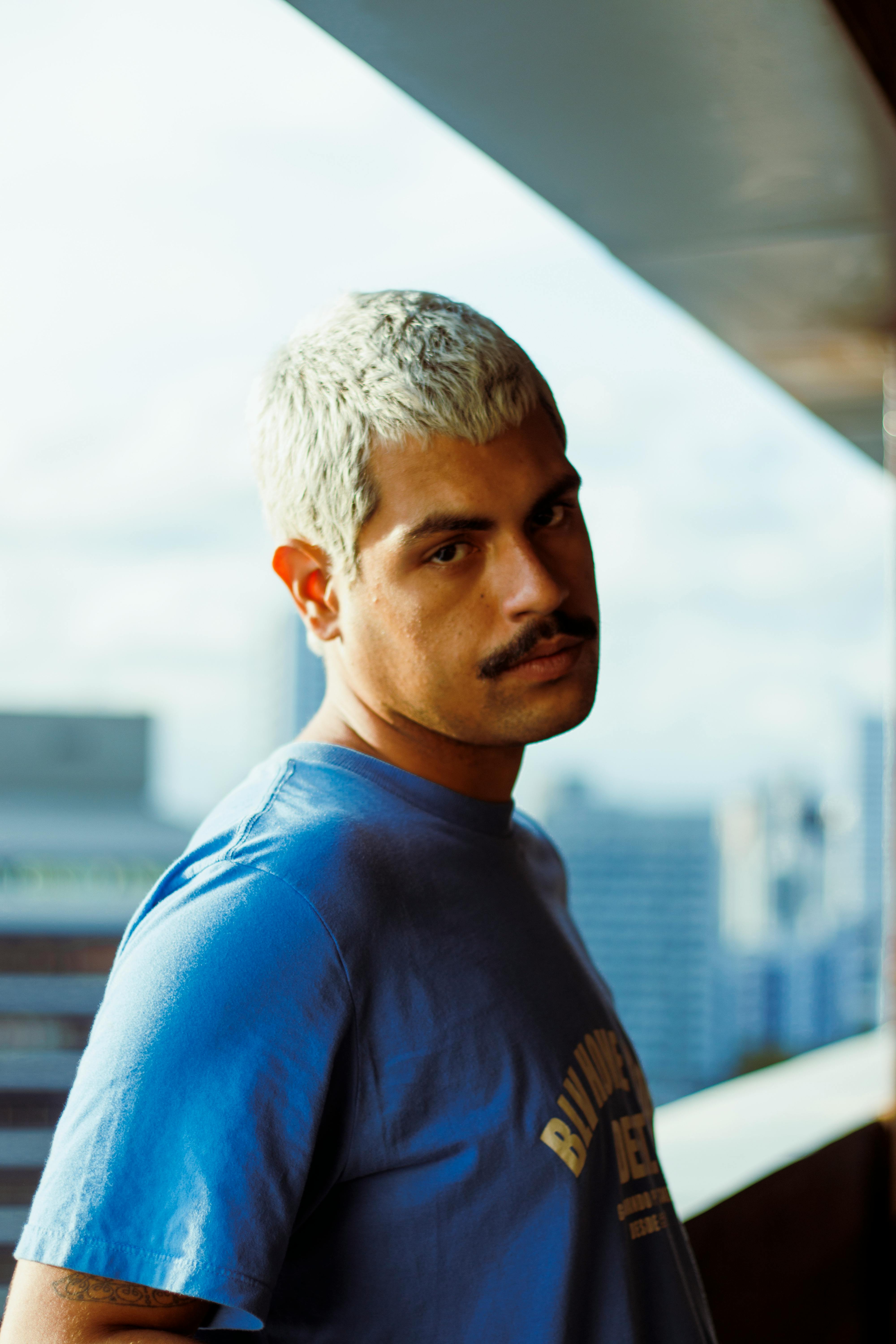 A man with blonde hair stands in an urban environment, wearing a blue shirt.