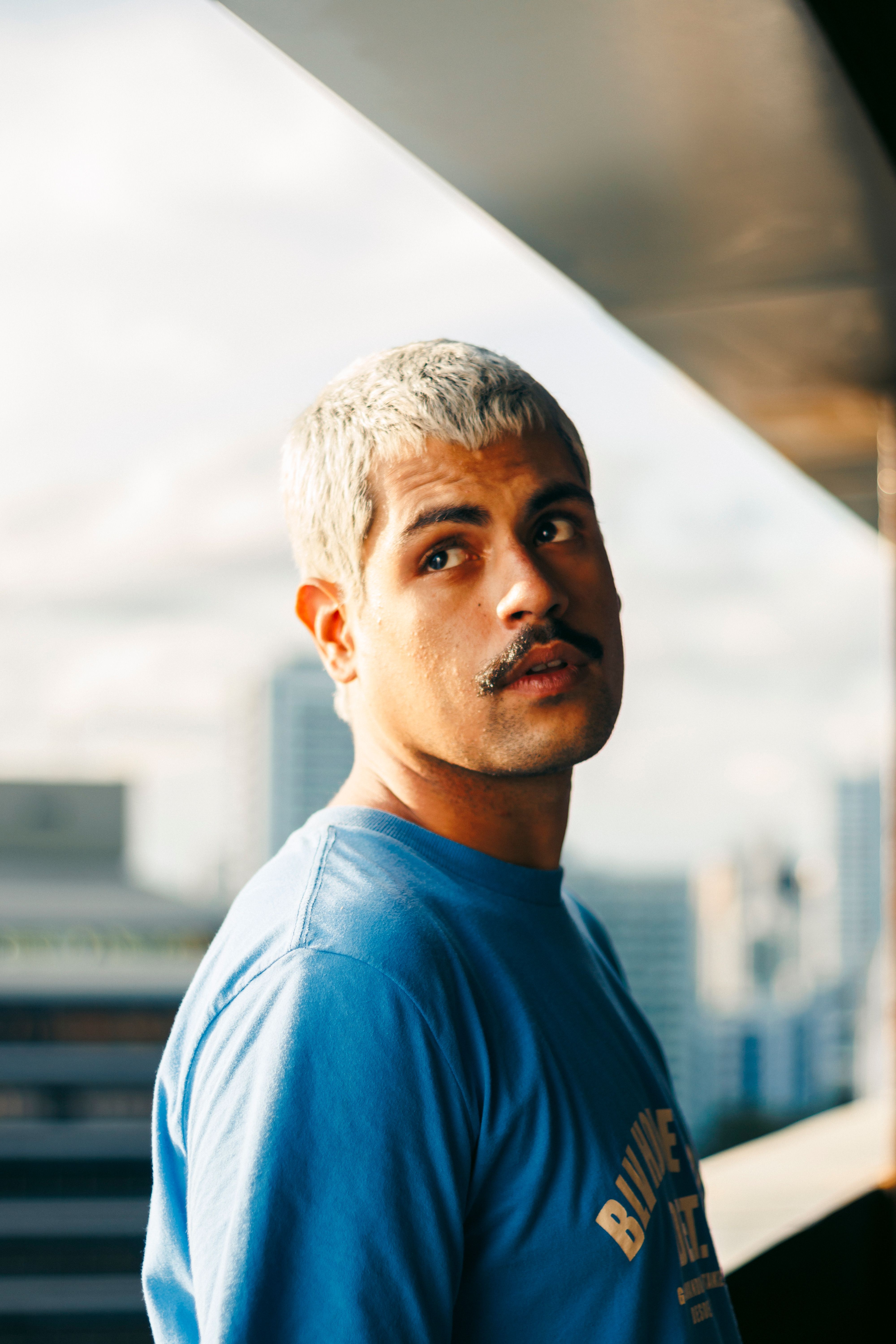 A man with a contemplative expression stands in an urban environment, bathed in natural light.