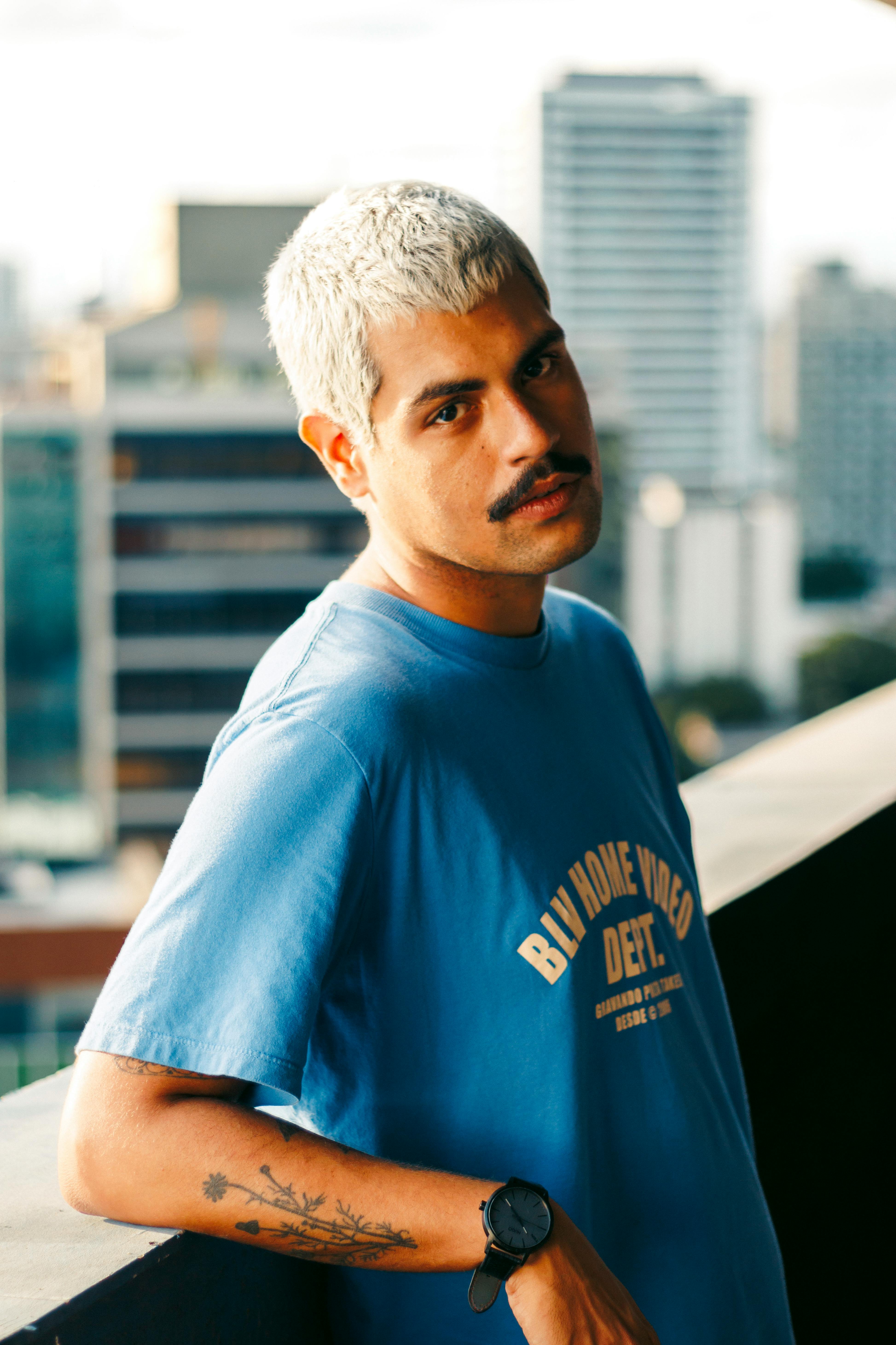 Stylish man with a mustache stands on a city balcony, wearing a blue shirt.