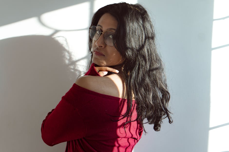 A woman in a red sweater poses gracefully with shadows on the wall in a sunlit room.