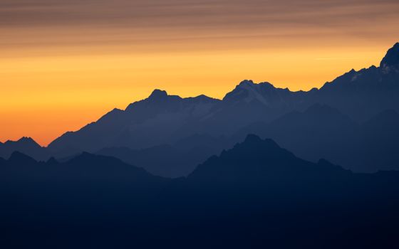 A breathtaking sunset over the Alpine mountain range with layered silhouettes and a warm, orange sky.