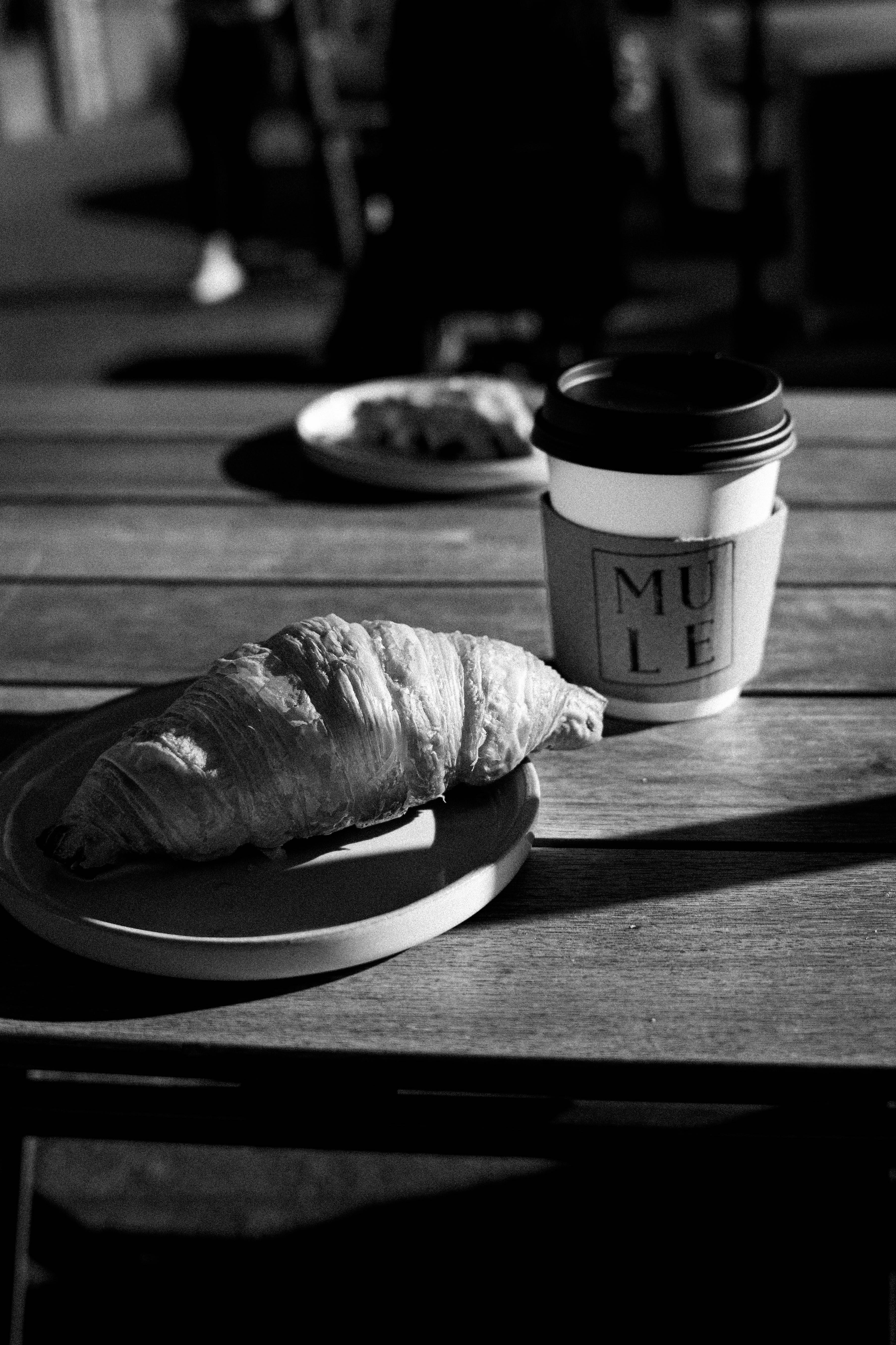 Artistic black and white photo of a croissant and coffee on a wooden table.