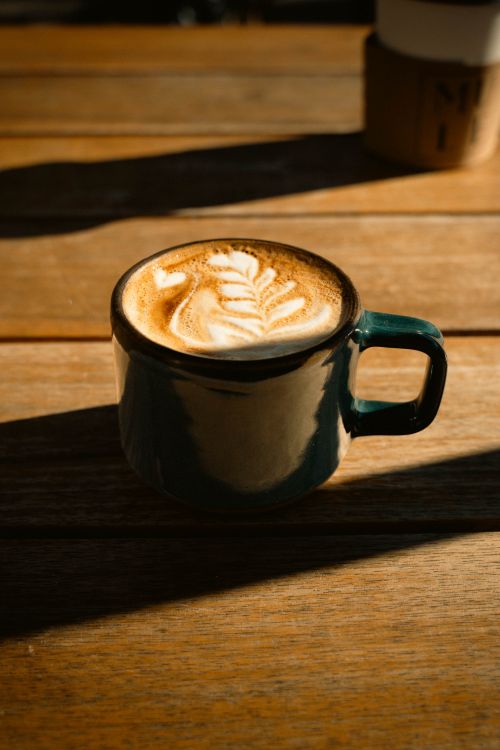 Free A beautiful cup of latte coffee with intricate art on a wooden table, basking in sunlight. Stock Photo