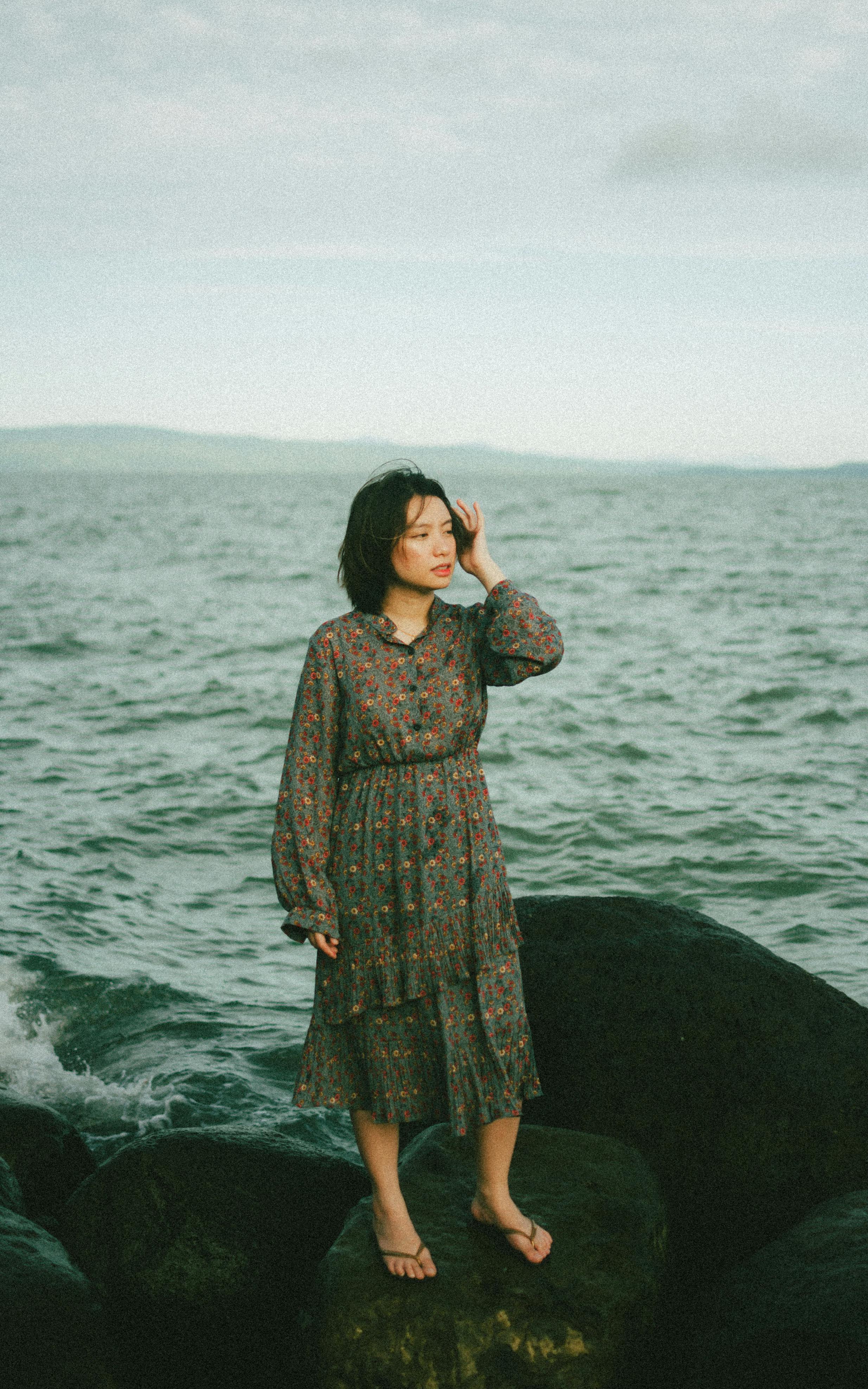 A woman in a floral dress stands on rocks by the sea, evoking tranquility.