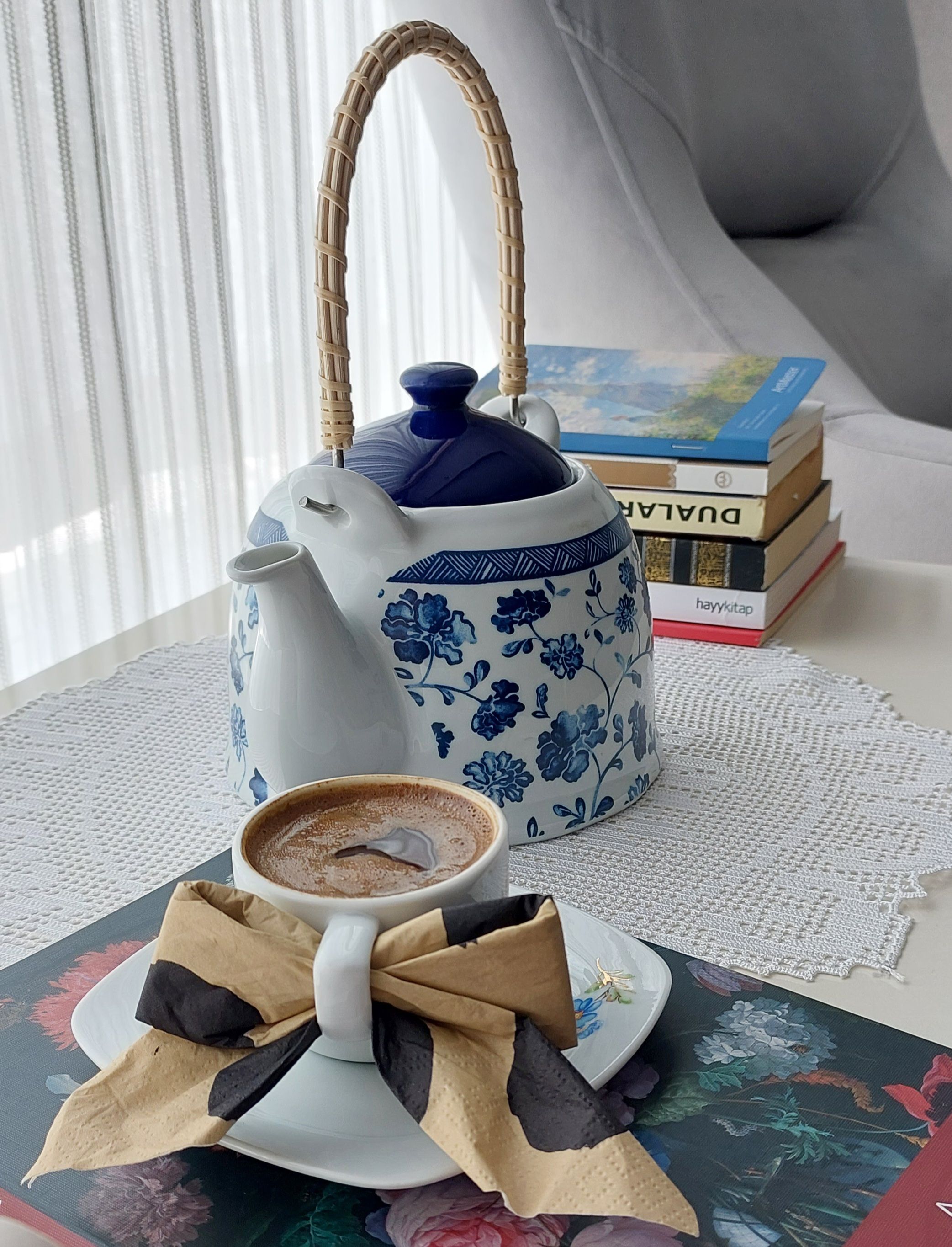 A cozy setup with a decorative teapot, a cup of coffee, and stacked books in a sunny indoor setting.