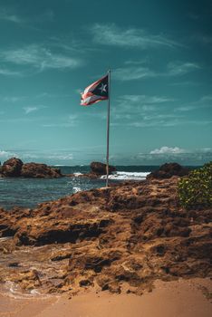 Puerto Rican flag waving on rocky coastal area in San Juan under a moody sky.