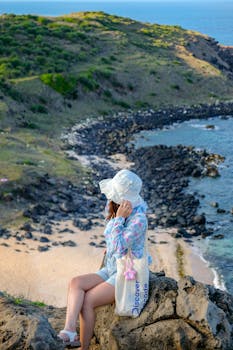 Woman in summer attire sitting on rocks overlooking a scenic beach view.