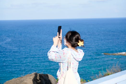 A woman taking a photo on a sunny day by the ocean with a smartphone.