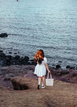 A young woman in a white dress carrying a bag walks along a rocky beach shore.
