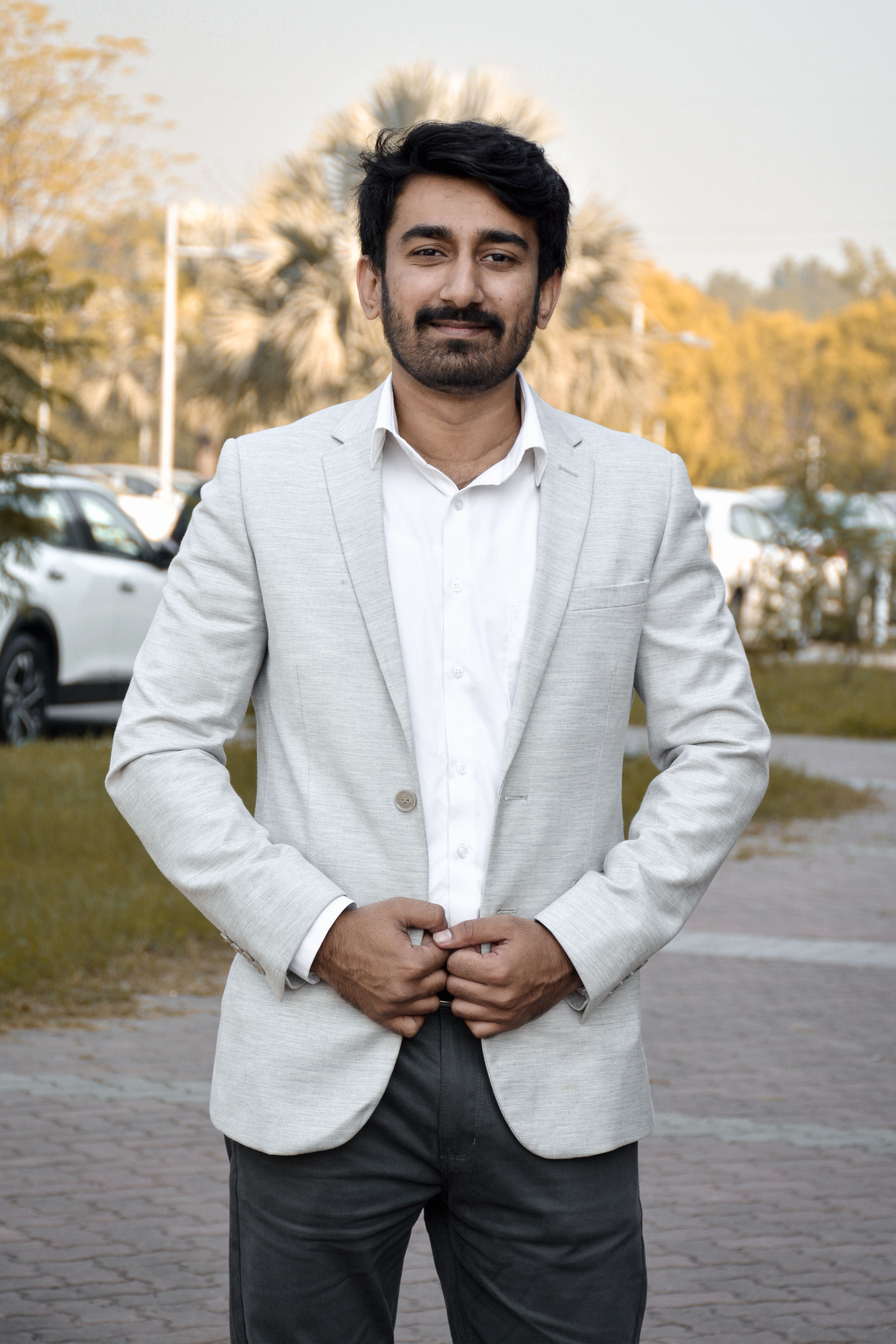 Confident man in white suit standing outdoors with cars and trees in the background.