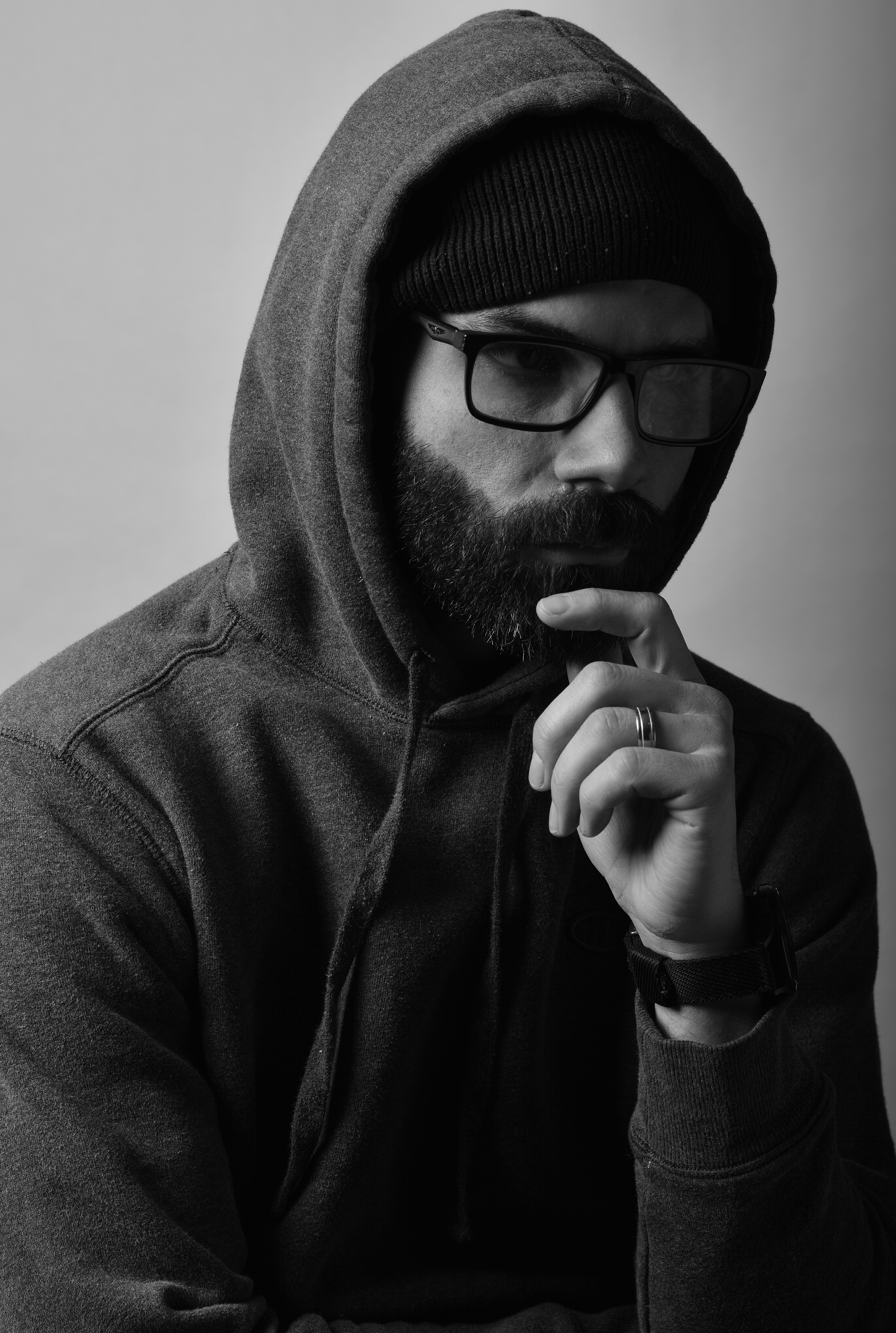 Black and white portrait of a bearded man in a hoodie and glasses deep in thought.