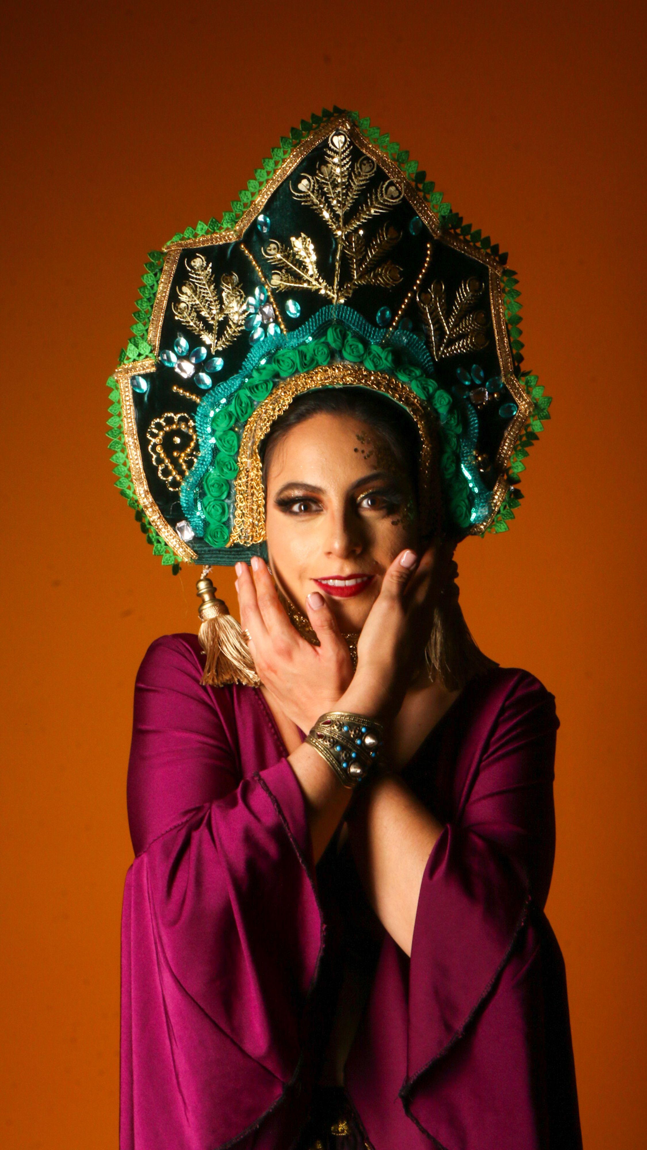 Woman wearing a vibrant traditional costume with an ornate headdress, posing against an orange background.