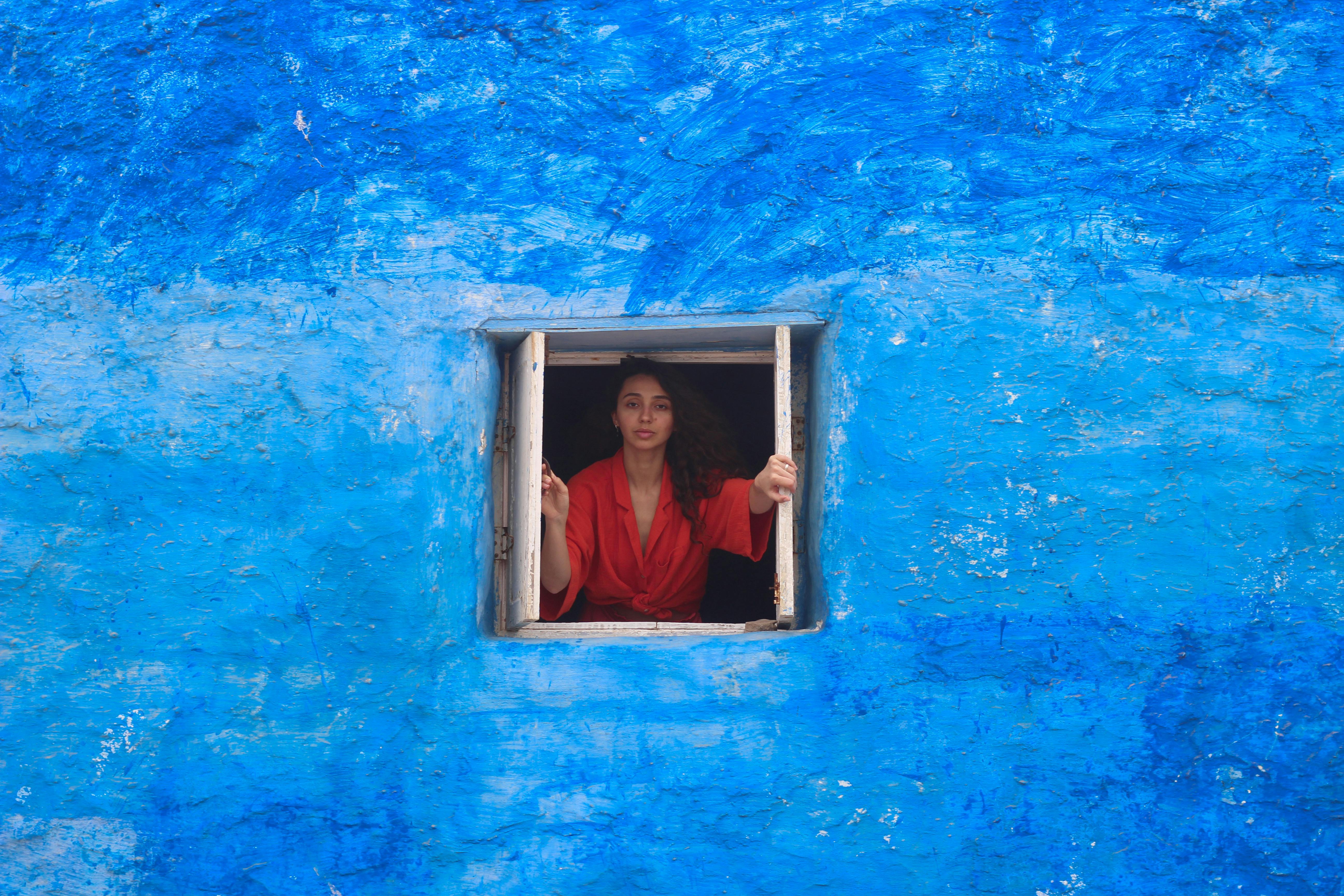 A woman in a red dress stands at a window in a blue wall in Casablanca, Morocco.