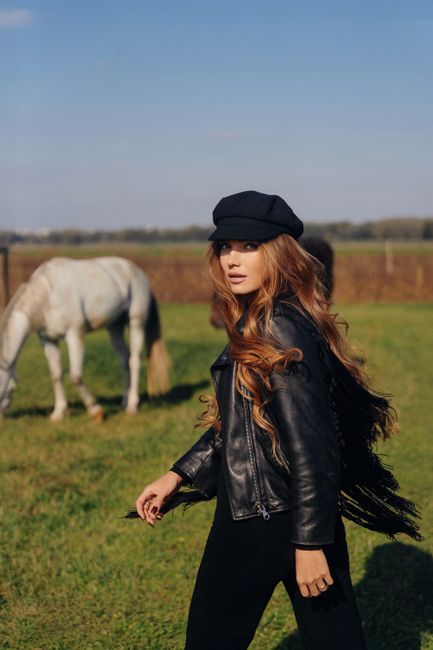 Stylish young woman in black leather jacket walking in a field with a white horse in the background.