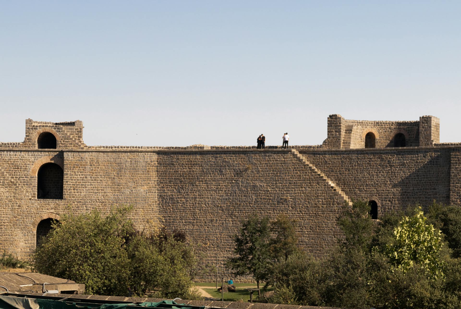 A historic stone fort with two people walking on top, surrounded by trees under a clear sky.