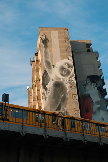 A Berlin street scene featuring striking building graffiti and passing train under clear skies.
