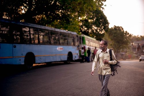 Free An adult man walks beside a bus on a tree-lined urban street during sunset, carrying a backpack. Stock Photo