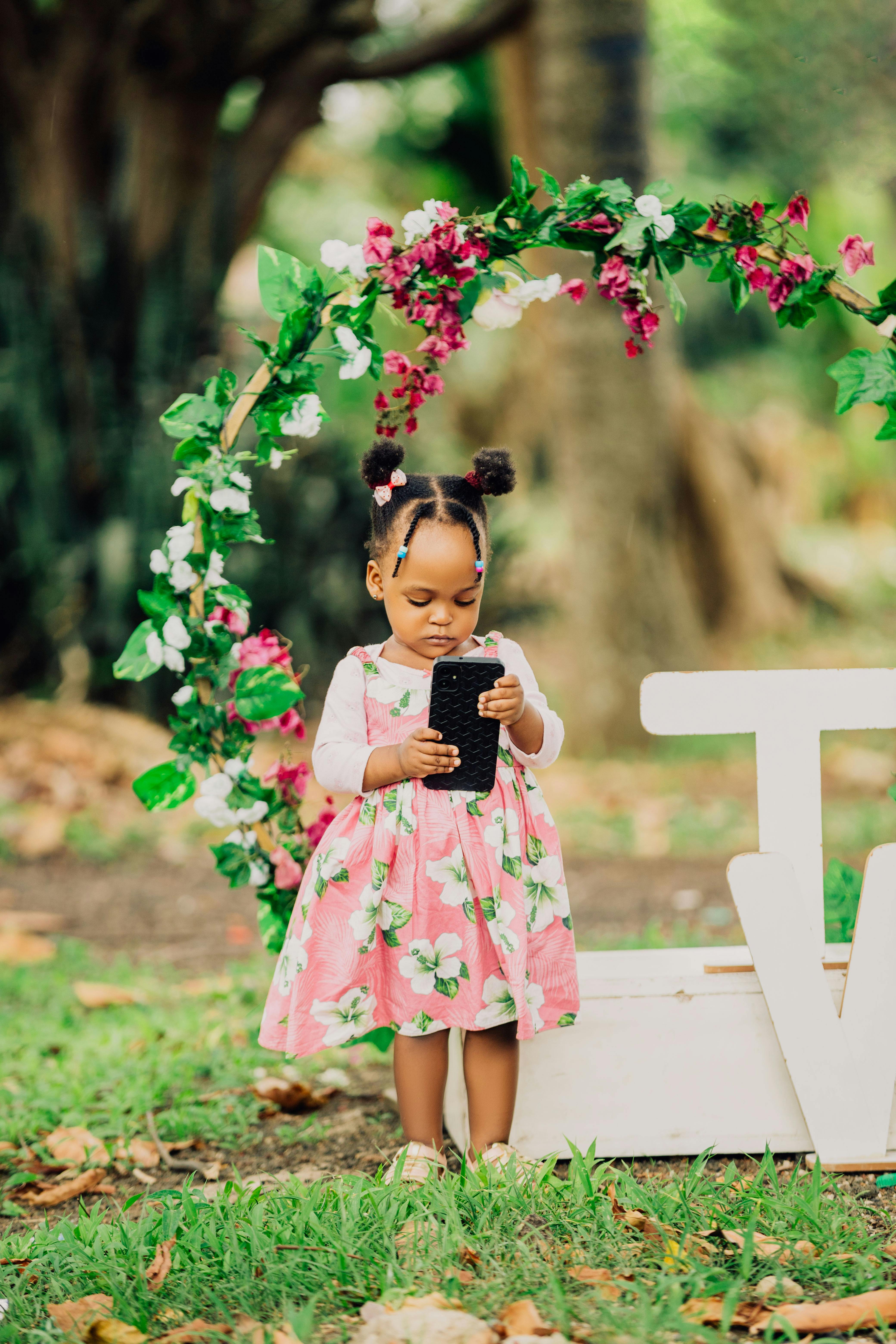 Gratis Gadis kecil dengan telepon pintar di taman dekat lengkungan bunga. Foto Stok